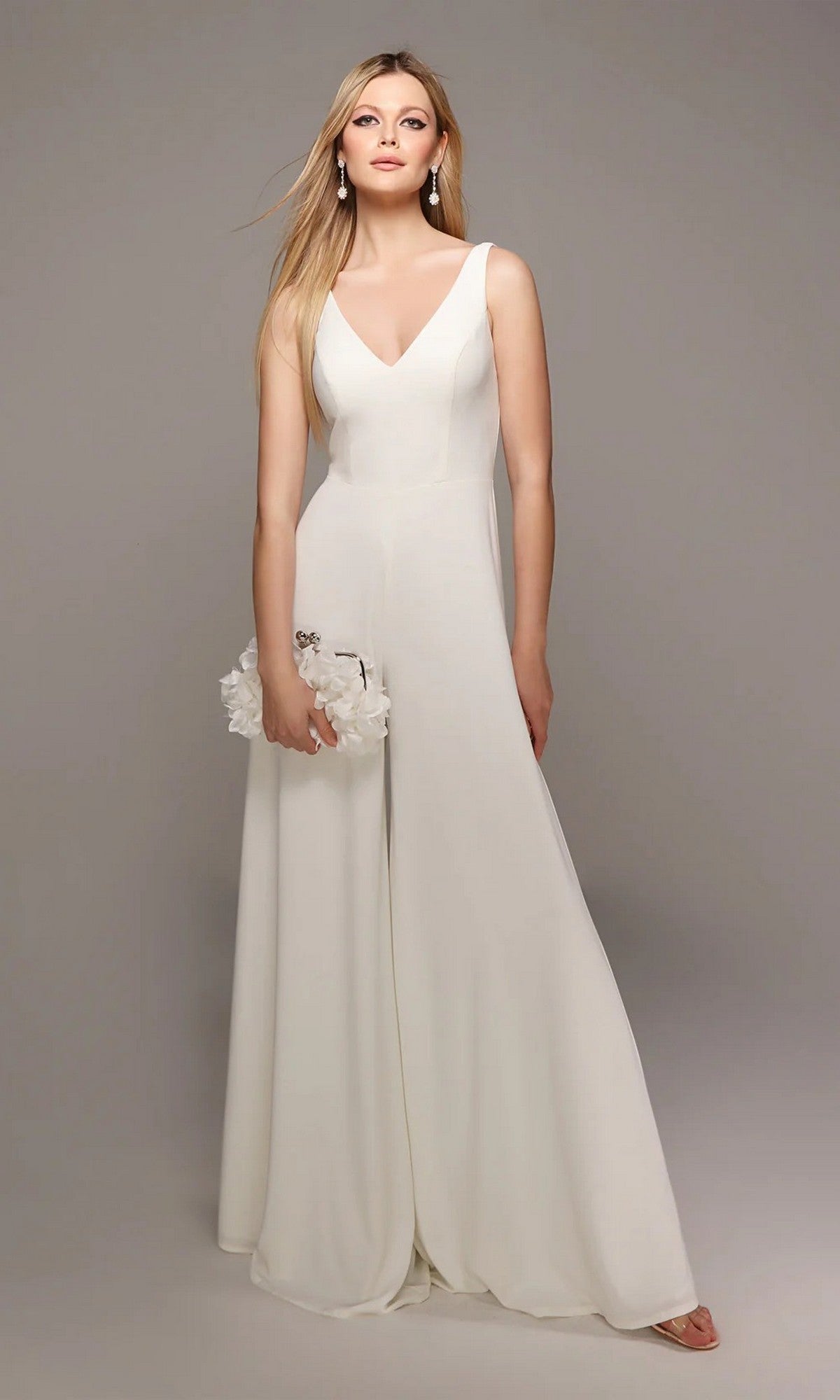 A woman models the Alyce 70021 long white jumpsuit in ivory, featuring wide-leg pants and a sleeveless design, holding a white clutch against a plain gray background.